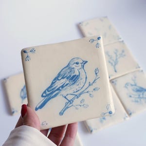 Könnte beinhalten: Eine quadratische Keramikfliese mit einer blauen Tuschezeichnung eines Vogels, der auf einem Ast sitzt. Die Fliese hat einen cremeweißen Hintergrund mit kleinen blauen Blattakzenten in den Ecken. Mehrere identische Fliesen sind im Hintergrund angeordnet.