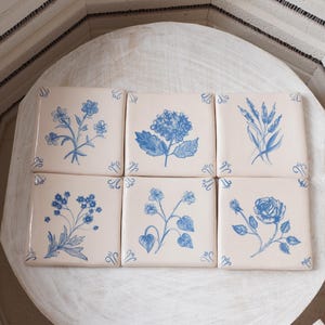 Pode incluir: Seis azulejos de cerâmica quadrados dispostos em duas filas de três. Cada azulejo de cor branco-sujo apresenta uma ilustração botânica azul distinta, incluindo várias flores e espigas de trigo, com pequenos detalhes decorativos azuis nos cantos.