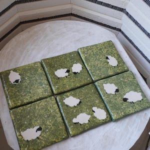 May include: A set of six square ceramic tiles featuring a glossy green speckled background with small white stars and illustrations of white sheep with black faces and legs. The tiles are arranged in a grid pattern on a circular white surface.
