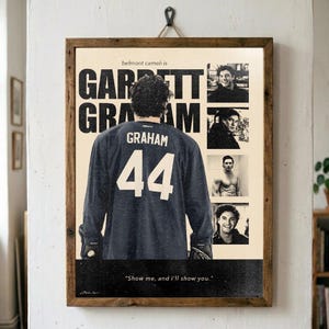 Puede incluir: Un póster rectangular en un marco de madera que muestra a un jugador de hockey con una camiseta oscura con el nombre GRAHAM y el número 44 en la espalda. El póster incluye el texto belmont cameli is GARRETT GRAHAM y Show me, and I'll show you.