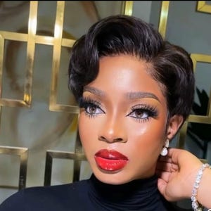 Peut inclure: Une femme aux cheveux noirs courts et ondulés, coiffés en coupe pixie. Elle porte un maquillage complet, avec de longs cils et un rouge à lèvres rouge vif. Elle porte un col roulé noir et des boucles d'oreilles en perles.