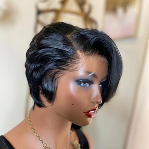 May include: A black, short-layered wig styled with a side part. The wig is on a mannequin head with blue eye shadow and red lipstick. A gold necklace is visible.