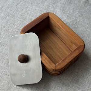 Puede incluir: Una mantequera de madera con forma rectangular y esquinas redondeadas. La tapa es de color plateado con un pequeño pomo redondo de madera. La mantequera está abierta, revelando la veta natural de la madera. La mantequera está sobre una superficie de lino de color neutro.