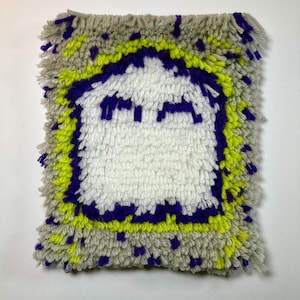 May include: A rectangular latch hook wall hanging featuring a central white ghost figure with purple eyes and a mouth. The ghost is surrounded by a bright neon yellow border, set against a textured light gray background with purple and yellow accents.