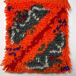 May include: A rectangular latch hook wall hanging featuring a vibrant orange background with two diagonal, textured grey and white abstract shapes. A bold red diagonal stripe cuts through the centre, accented by small purple yarn details.