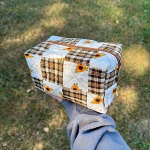 May include: A rectangular quilted zippered pouch featuring a patchwork design of alternating brown and white plaid squares and white squares decorated with small yellow sunflower illustrations. The bag has a brown zipper closure along the top edge.