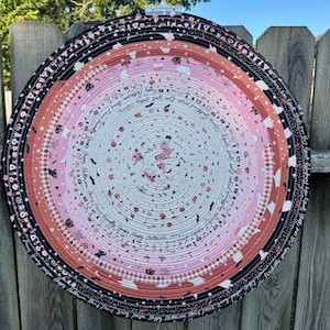 May include: A circular fabric mat constructed from coiled rope with a gradient colour scheme transitioning from white at the centre to pink, orange, and black at the edge. The fabric features Halloween-themed prints including ghosts, cats, and text.