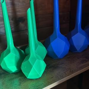 May include: A collection of 3D-printed objects with a geometric design. The objects are in shades of green and blue, with a faceted, bulbous base and a long, slender spout. They are arranged on a wooden shelf.