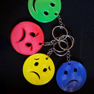 May include: A set of four colorful keychains featuring smiley and sad faces. The keychains are round and made of plastic, with a green happy face, a red sad face, a yellow sad face, and a blue sad face. Each keychain is attached to a metal ring.