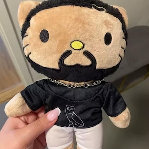 May include: A plush toy in the style of Hello Kitty, featuring a brown face, black beard, and a black and gold chain necklace. It is dressed in a black shirt with a white owl logo and white trousers. The toy has a yellow nose and black eyes.