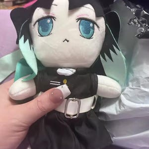 May include: A plush doll with large blue eyes and black hair. The doll is wearing a black outfit with a white belt and a small gold button. The doll has a white face and small black details. The doll has mint green accents.