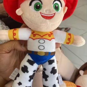 May include: A plush doll of a character with a red hat and red hair. The doll has a white shirt with yellow and red accents, blue pants, and white legs with black spots. The doll has green eyes and a smiling expression.