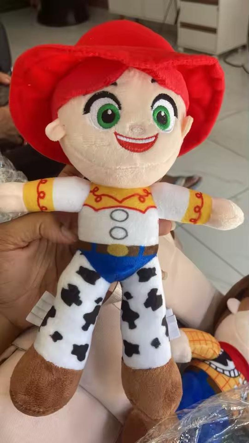 May include: A plush doll of a character with a red hat and red hair. The doll has a white shirt with yellow and red accents, blue pants, and white legs with black spots. The doll has green eyes and a smiling expression.