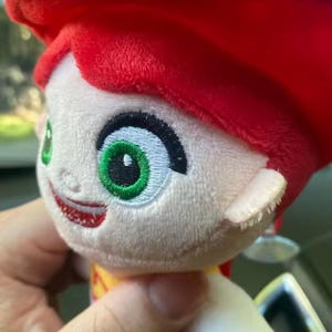 May include: A plush doll of a character with red hair, green eyes, and a smiling expression. The doll has a red hat and a yellow and red outfit. The doll is held by a person.