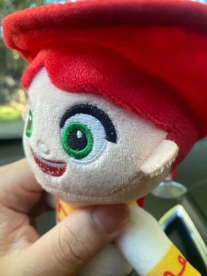 May include: A plush doll of a character with red hair, green eyes, and a smiling expression. The doll has a red hat and a yellow and red outfit. The doll is held by a person.