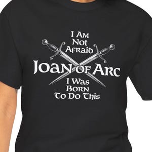 May include: A black t-shirt featuring a white graphic of two crossed swords. The text reads: I AM NOT AFRAID JOAN OF ARC I WAS BORN TO DO THIS. The design is centered on the chest, utilizing a bold, serif-style font for the lettering.