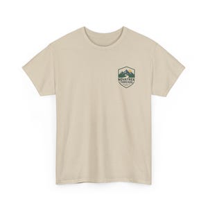 Könnte beinhalten: Beiges T-Shirt mit kleinem Aufdruck auf der linken Brust. Die Grafik zeigt eine Bergkette, eine Sonne und den Text "NOVATREK THREADS". Das Shirt ist aus weichem Material und hat einen Rundhalsausschnitt.