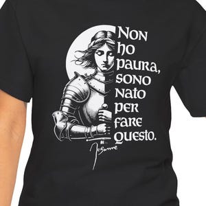 Könnte beinhalten: Ein schwarzes T-Shirt mit einer weißen grafischen Illustration einer Figur in mittelalterlicher Rüstung. Der Text lautet: "NON ho paura, SONO Nato peR faRe questo." und enthält eine Signatur, die "Jeanne" lautet, am unteren Rand.