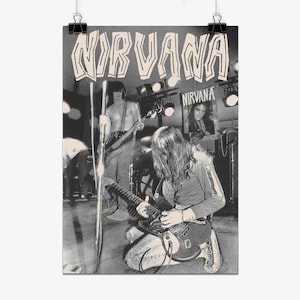 May include: A black and white poster featuring the band Nirvana. The image shows a musician playing an electric guitar while kneeling on stage, with another band member in the background. The word NIRVANA is printed in large, stylized letters at the top.
