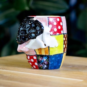 Pode incluir: Uma caneca de cerâmica colorida com um design único. A caneca apresenta um padrão de patchwork com bolinhas vermelhas, detalhes amarelos, azuis e florais. Um rosto preto texturizado com bigode e nariz brancos é esculpido na lateral.