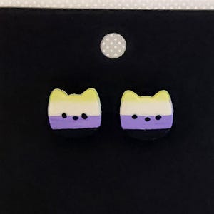 May include: A pair of stud earrings shaped like cat heads. Each earring features horizontal stripes of yellow, white, purple, and black, mimicking the non-binary pride flag colors. The cat faces have two small black dots for eyes and a black nose.