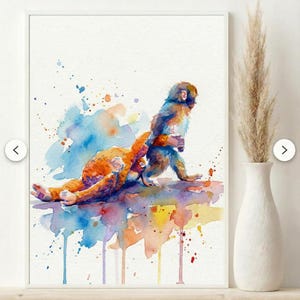 May include: Watercolour painting of two monkeys in shades of orange, blue, and brown. The artwork features colourful watercolour splashes and drips, set against a white background. The painting is framed in a white frame and displayed on a shelf.