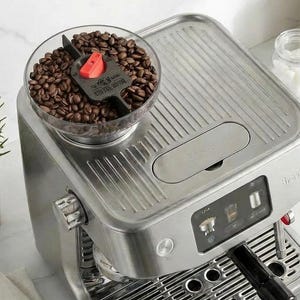 May include: A brushed stainless steel espresso machine with a top-mounted clear bean hopper filled with dark roasted coffee beans, a digital touchscreen interface, a black portafilter handle, and a ribbed drip tray base for brewing coffee.