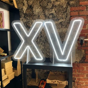Puede incluir: Un letrero de neón grande e iluminado que muestra las letras X y V en una luz blanca brillante. Las letras están montadas sobre soportes de acrílico transparente, cada una sostenida por una pequeña base de acrílico, creando una pieza decorativa.