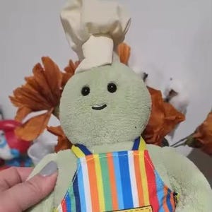 May include: A plush green turtle toy wearing a chef's hat and a colorful striped apron. The toy has a smiling face and is designed to look like a chef. The toy is soft and cuddly.