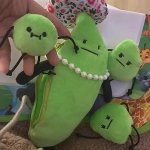 May include: A plush green pea pod with a pearl necklace, surrounded by three smaller pea plushies. Each plush has a stitched face and black string arms and legs. A floral patterned fabric is visible in the background.