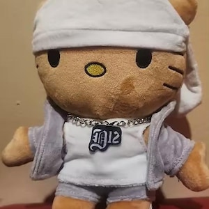 May include: A plush Hello Kitty doll wearing a grey tracksuit, white undershirt, and a white cap. The doll has a silver chain necklace and a pendant with the letters "J12". The doll is tan and brown with black eyes and whiskers.