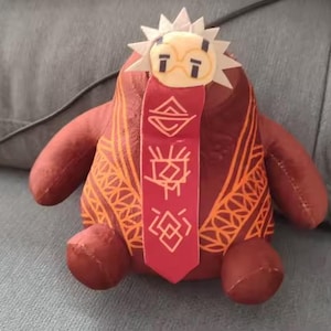 Könnte beinhalten: Ein Plüschpuppe in Rot- und Orangetönen, mit einem sonnenförmigen Kopfschmuck und einem roten Band mit weißen Symbolen. Die Puppe hat ein gemustertes Design auf ihrem Körper und ein lächelndes Gesicht.