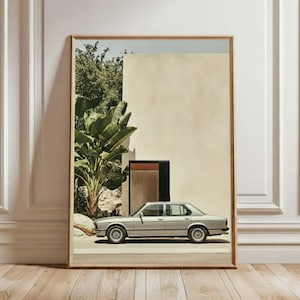 May include: A framed print showing a silver saloon car parked on a road. The car is in front of a beige wall, a dark door, and green foliage. The print is in a light brown wooden frame, leaning against a white wall and a wooden floor.