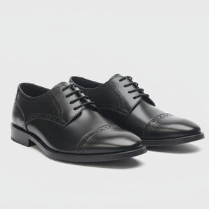 May include: A pair of black leather Oxford shoes with a cap toe and decorative stitching. The shoes have a lace-up closure and a low heel. The shoes are displayed on a white background.