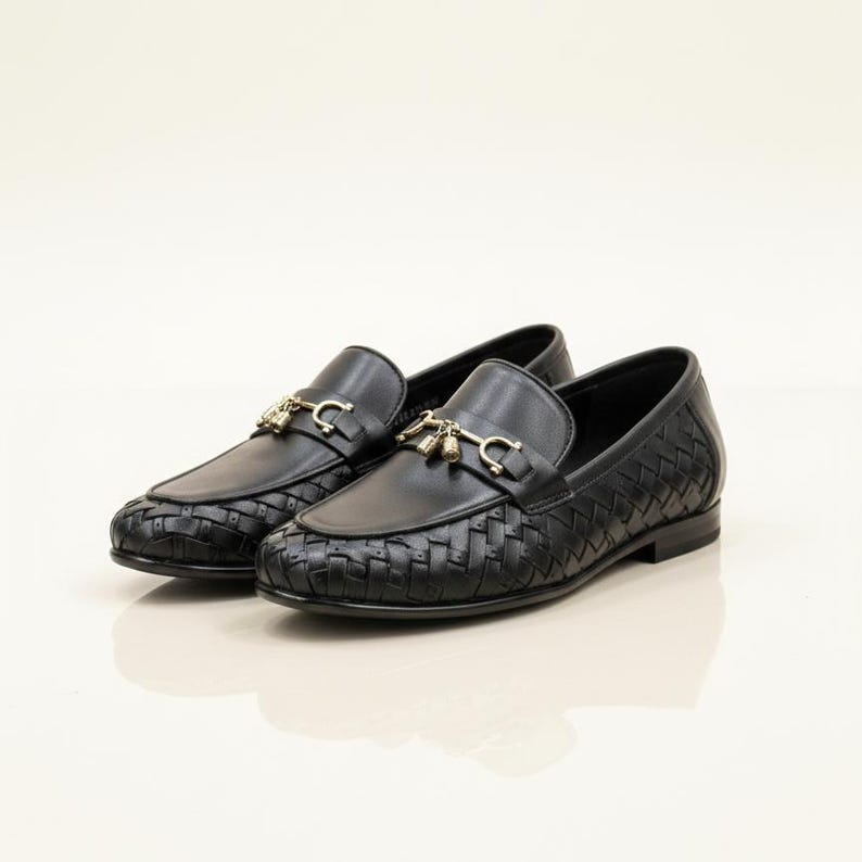 May include: A pair of black leather loafers with a woven design on the toe and sides. The shoes feature gold-colored metal accents on the top. The loafers are displayed on a white surface.
