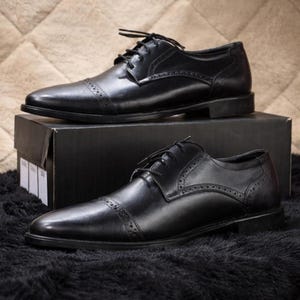May include: Two pairs of black leather dress shoes with cap toes and lace-up closures. The shoes are displayed on a black box and a black textured surface. The shoes have decorative perforations along the seams.