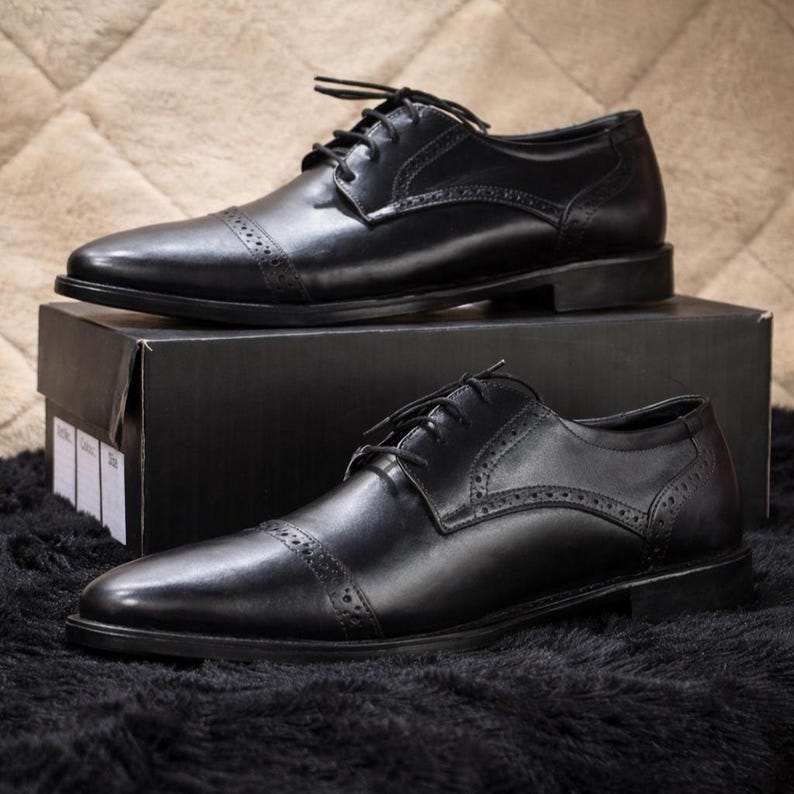 May include: Two pairs of black leather dress shoes with cap toes and lace-up closures. The shoes are displayed on a black box and a black textured surface. The shoes have decorative perforations along the seams.