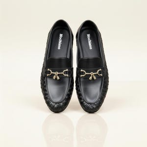 May include: A pair of black Mochiano loafers with a woven texture. Each shoe features a gold-colored metal accent with a decorative tassel. The brand name "Mochiano" is printed in white on the insole.