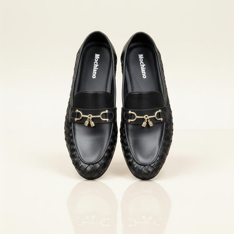 May include: A pair of black Mochiano loafers with a woven texture. Each shoe features a gold-colored metal accent with a decorative tassel. The brand name "Mochiano" is printed in white on the insole.