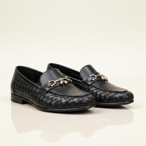 May include: A pair of black leather loafers with a woven texture on the vamp and sides. The shoes feature a gold-colored metal ornament on the upper. The loafers are displayed on a light-colored surface.