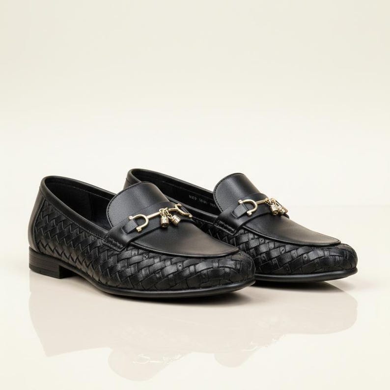 May include: A pair of black leather loafers with a woven texture on the vamp and sides. The shoes feature a gold-colored metal ornament on the upper. The loafers are displayed on a light-colored surface.