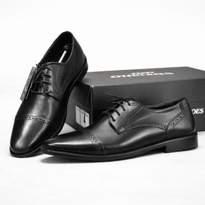 May include: Black leather dress shoes with a classic cap-toe design. The shoes have a lace-up closure and decorative perforations. A black shoebox is in the background. The shoes are a formal style.
