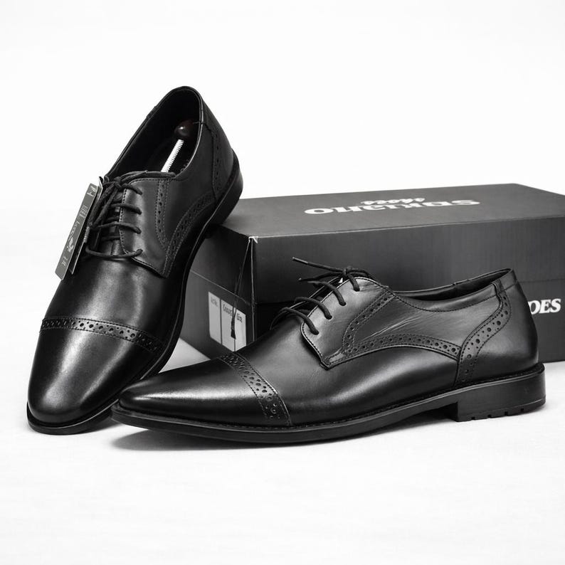 May include: Black leather dress shoes with a classic cap-toe design. The shoes have a lace-up closure and decorative perforations. A black shoebox is in the background. The shoes are a formal style.