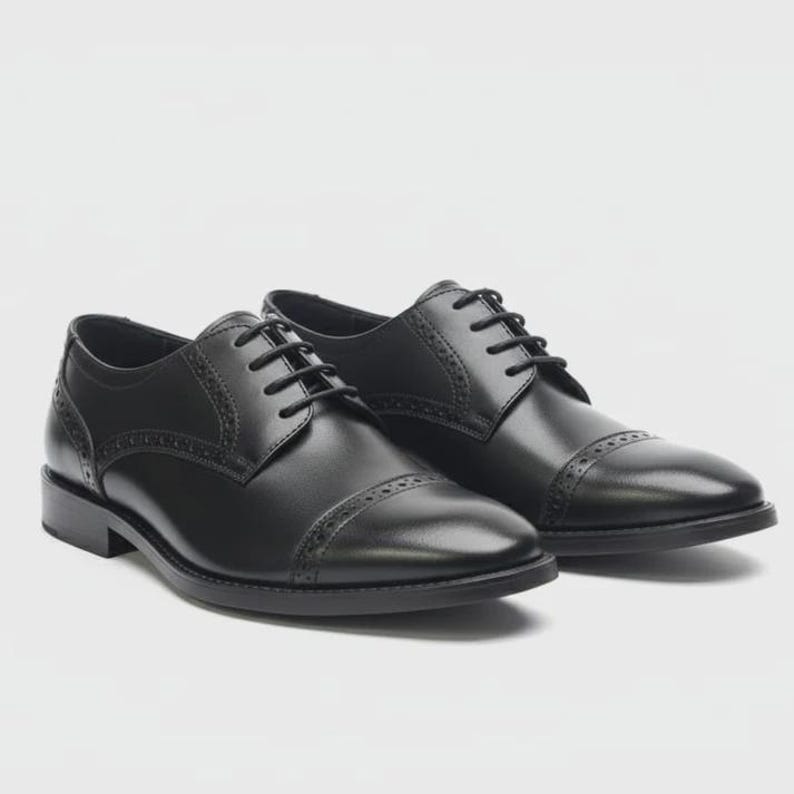 May include: A pair of black leather Oxford shoes with a cap toe and decorative stitching. The shoes have a lace-up closure and a low heel. The shoes are displayed on a white background.