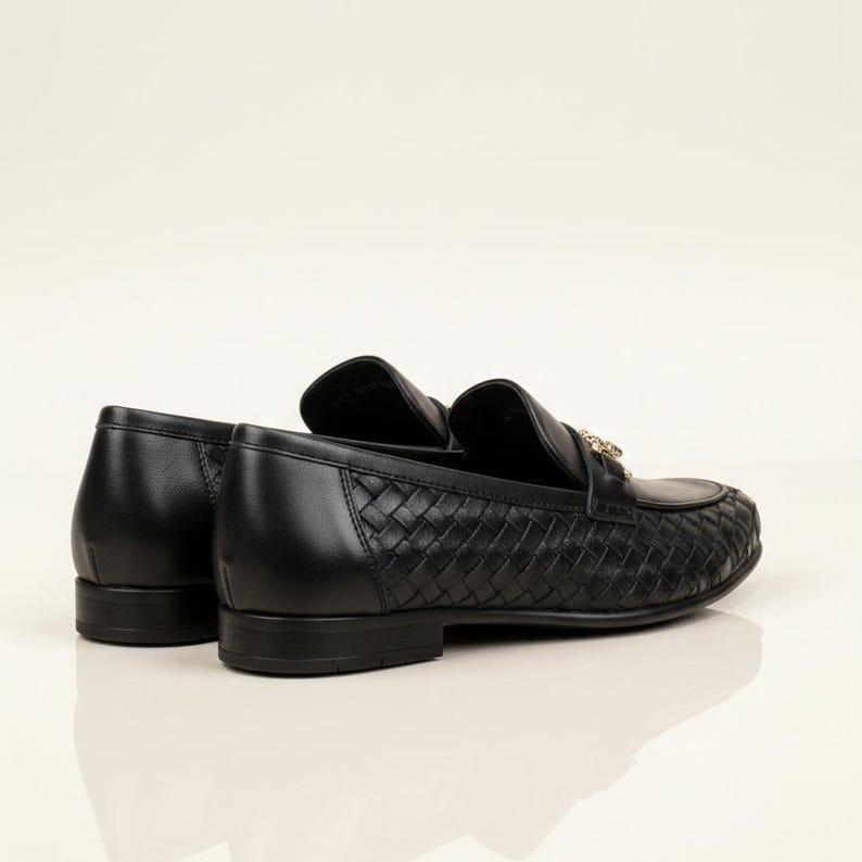 May include: Black leather loafers with a woven pattern on the vamp. The shoes have a gold-colored accent on the top and a low heel. The loafers are displayed on a white surface.
