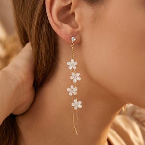 May include: A long gold-tone drop earring featuring a small clear crystal stud at the top, followed by a delicate gold chain adorned with four white five-petalled flower charms, each centred with a tiny clear crystal accent for a layered look.