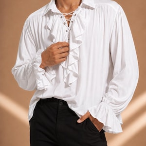 May include: A white long-sleeved shirt featuring a ruffled front placket, a lace-up neckline with silver-toned metal grommets, and gathered cuffs with ruffled edges. The shirt is paired with solid black trousers, creating a high-contrast look.