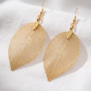 May include: A pair of gold-coloured leaf-shaped earrings. The earrings have a delicate, veined design, with a small leaf accent at the top. The earrings hang from gold-coloured ear wires.