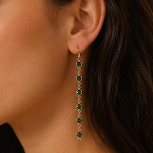 May include: A long gold drop earring featuring a delicate chain design with six evenly spaced, circular green stones set in gold bezels. The earring hangs vertically from a simple gold hook, creating a linear silhouette with vibrant green accents.