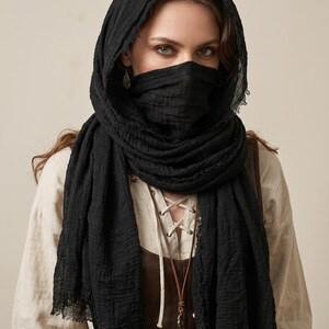 May include: A black textured scarf draped over the head and wrapped around the lower face, featuring frayed edges and a lightweight, crinkled fabric construction. The scarf is worn over a beige long-sleeve shirt and a brown leather-like corset top.